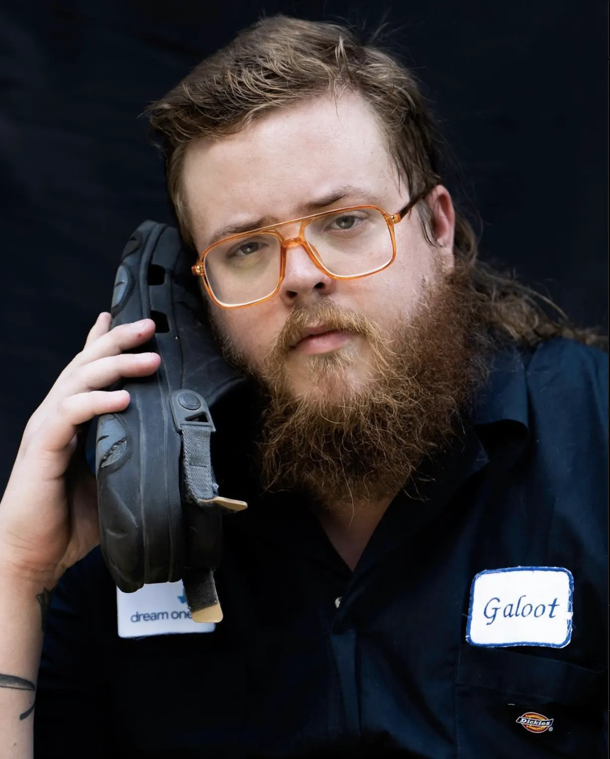 Zachary Boyd wearing orange glasses and a Galoot work shirt, holding a sandal to his ear like a telephone